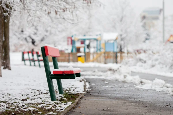 Stock image Red bench in a snowy park, children playground in the background