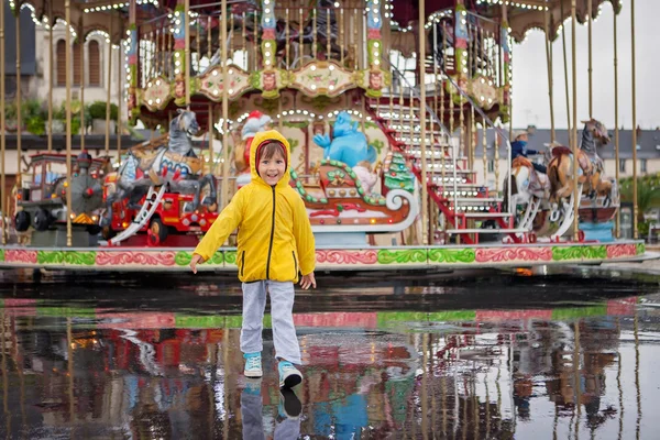 Sweet child, boy watching carousel in the rain, wearing yellow r — Zdjęcie stockowe