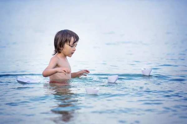 Ένα μικρό χαριτωμένο αγόρι στην παραλία με χάρτινες βάρκες. Ευτυχισμένα παιδιά PL — Φωτογραφία Αρχείου