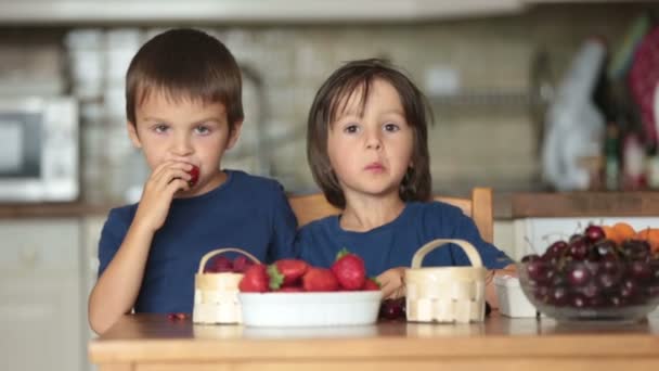 Két édes gyermekeim, fiú testvér, eszik otthon friss gyümölcsök, eper, cseresznye, málna, áfonya — Stock videók