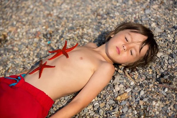 Schattige kleine jongen, liggend in het zand op het strand, twee rode sta — Stockfoto