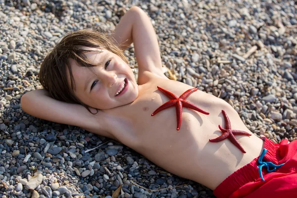 Αξιολάτρευτο μικρό αγόρι, ξαπλωμένοι στην άμμο στην παραλία, δύο κόκκινα sta — Φωτογραφία Αρχείου