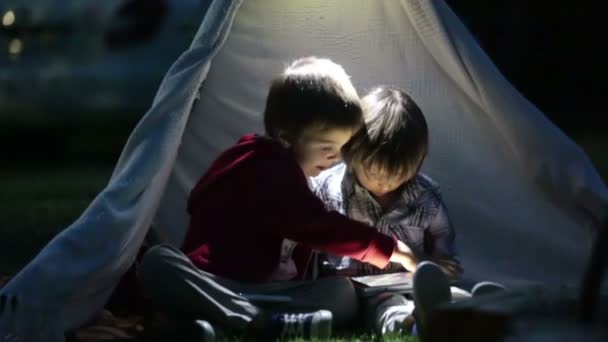 Lindos hermanos pequeños, jugando en la tableta y el teléfono por la noche en el campamento, en la tienda de campaña — Vídeos de Stock