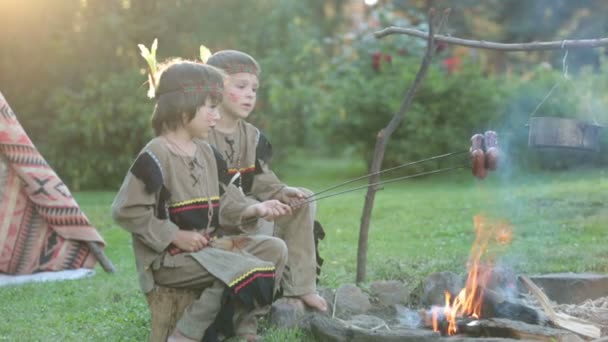 Симпатичный портрет коренных американцев в костюмах, играющих на свежем воздухе вокруг костра на закате, готовящих сосиски, летом — стоковое видео