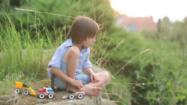 Menino bonito, brincando com pequenos carros de brinquedo na rua ao pôr-do-sol, horário de verão, de volta iluminado — Vídeo de Stock