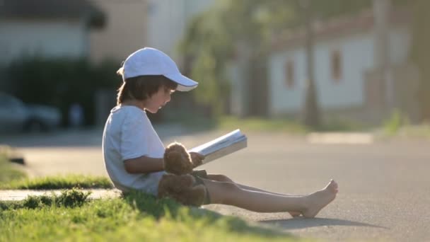 Beautiful kid boy, reading a book on the street, sitting down with teddy bear, gorgeous sunset light — Stock Video