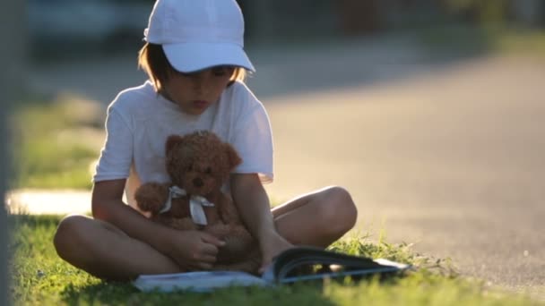 Красивый мальчик, читает книгу на улице, сидит с плюшевым мишкой, великолепный закат света — стоковое видео