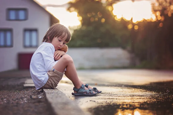 Triste niño, sentado en la calle bajo la lluvia, abrazando su t — Foto de Stock