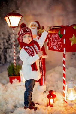 Küçük bir çocuk, küçük bir çocuk, Noel Baba 'ya posta kutusuyla mektup yolluyor, etrafında Noel süsü, açık havada karla vuruyor.