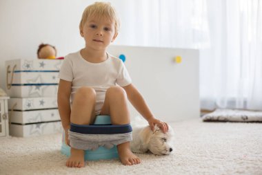 Sarışın çocuk, evde lazımlık kullanıyor, yanında evcil bir köpek yatıyor.