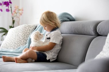 Sevimli küçük sarışın çocuk, yeni yürümeye başlayan çocuk, evde beyaz köpek yavrusuyla oynuyor.