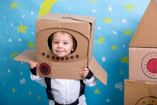 Sweet toddler boy, dressed as an astronaut, playing at home with cardboard rocket and handmade helmet from box