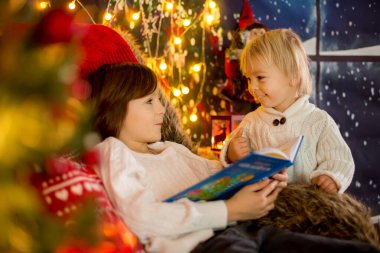 Şirin çocuk ve ağabeyi, Noel ağacının etrafında rahat bir sandalyeye oturmuş, Noel gecesi kitap okuyorlar.