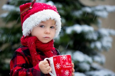 Tatlı güzel çocuk, elinde sıcak sütle bardağı tutuyor, karda dışarıda içiyor, kışın ve Noel tatilinin tadını çıkarıyor.