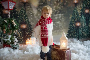 Elinde bavuluyla küçük bir çocuk ve elinde küçük bir kızıladam oyuncağı, karlı bir ormanda yürüyor, Noel temalı bir resim.