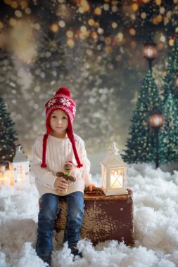 Elinde bavuluyla küçük bir çocuk ve elinde küçük bir kızıladam oyuncağı, karlı bir ormanda yürüyor, Noel temalı bir resim.