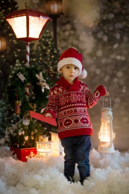 Tatlı sarışın çocuk, kırmızı kazaklı, ormanda fener tutan, Noel zamanı, kar yağıyor.