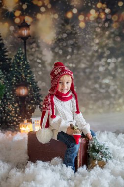 Elinde bavuluyla küçük bir çocuk ve elinde küçük bir kızıladam oyuncağı, karlı bir ormanda yürüyor, Noel temalı bir resim.