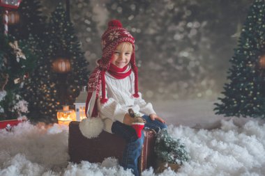 Elinde bavuluyla küçük bir çocuk ve elinde küçük bir kızıladam oyuncağı, karlı bir ormanda yürüyor, Noel temalı bir resim.