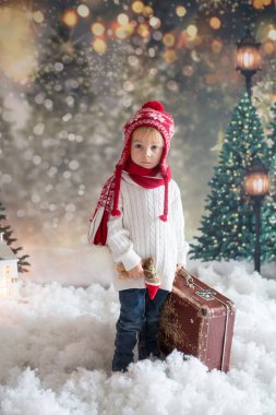 Elinde bavuluyla küçük bir çocuk ve elinde küçük bir kızıladam oyuncağı, karlı bir ormanda yürüyor, Noel temalı bir resim.