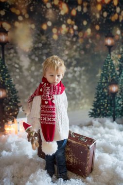 Elinde bavuluyla küçük bir çocuk ve elinde küçük bir kızıladam oyuncağı, karlı bir ormanda yürüyor, Noel temalı bir resim.