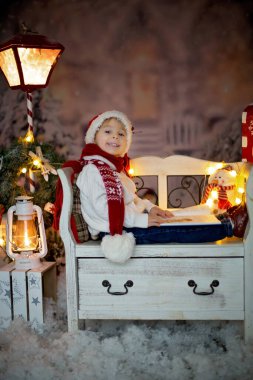 Güzel bir çocuk, sarışın bir çocuk, kitap okuyor, bir bankta oturuyor, etrafında Noel süslemeleri var.