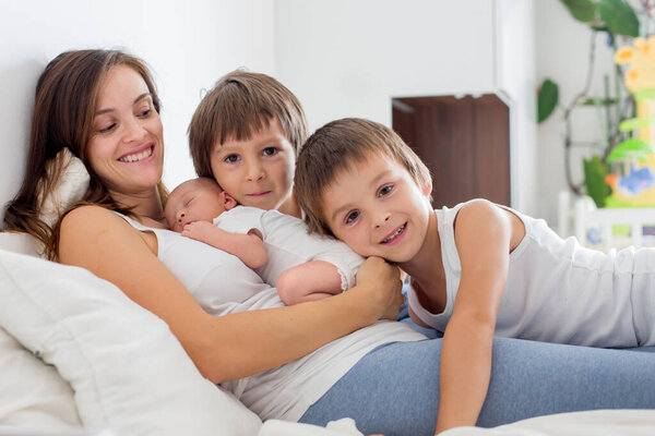 Young mother lying in bed with her newborn baby boy and his older brothers, holding them in her arms and smiling from happines