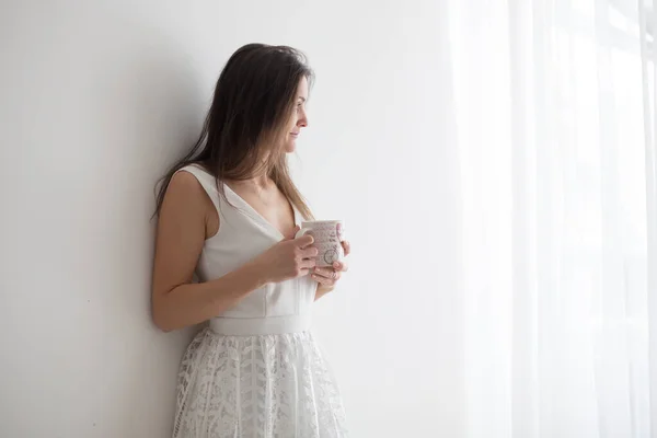 白いドレスを着た中年女性家でコーヒーを飲み窓から外を見る白い背景 — ストック写真