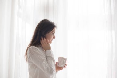 Beyaz elbiseli orta yaşlı bir kadın. Evde kahve içiyor. Pencereden dışarı bakıyor.