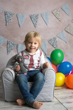 Şirin sarışın çocuk, çocuk, küçük bebek koltuğunda oturuyor, renkli balonlarla oynuyor, mutlu bir şekilde gülümsüyor, kapalı alanda fotoğraf çekiyor.