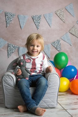 Şirin sarışın çocuk, çocuk, küçük bebek koltuğunda oturuyor, renkli balonlarla oynuyor, mutlu bir şekilde gülümsüyor, kapalı alanda fotoğraf çekiyor.