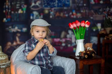 Tatlı çocuk, gözlük takıyor, kitap okuyor, kurabiyelerle çay içiyor, rahat bir sandalyede oturuyor.