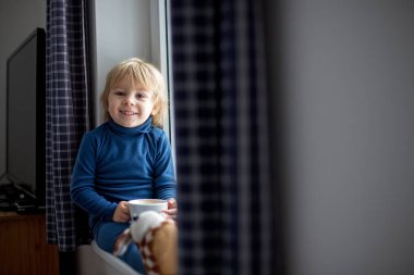 Küçük çocuk, pencerede oturuyor, karın düşüşünü izliyor, bir fincan çayın tadını çıkarıyor.