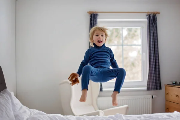 幸せな幼児の子供 青いパジャマのブロンドの男の子 ベッドの上でジャンプ 小さなおもちゃを保持 — ストック写真