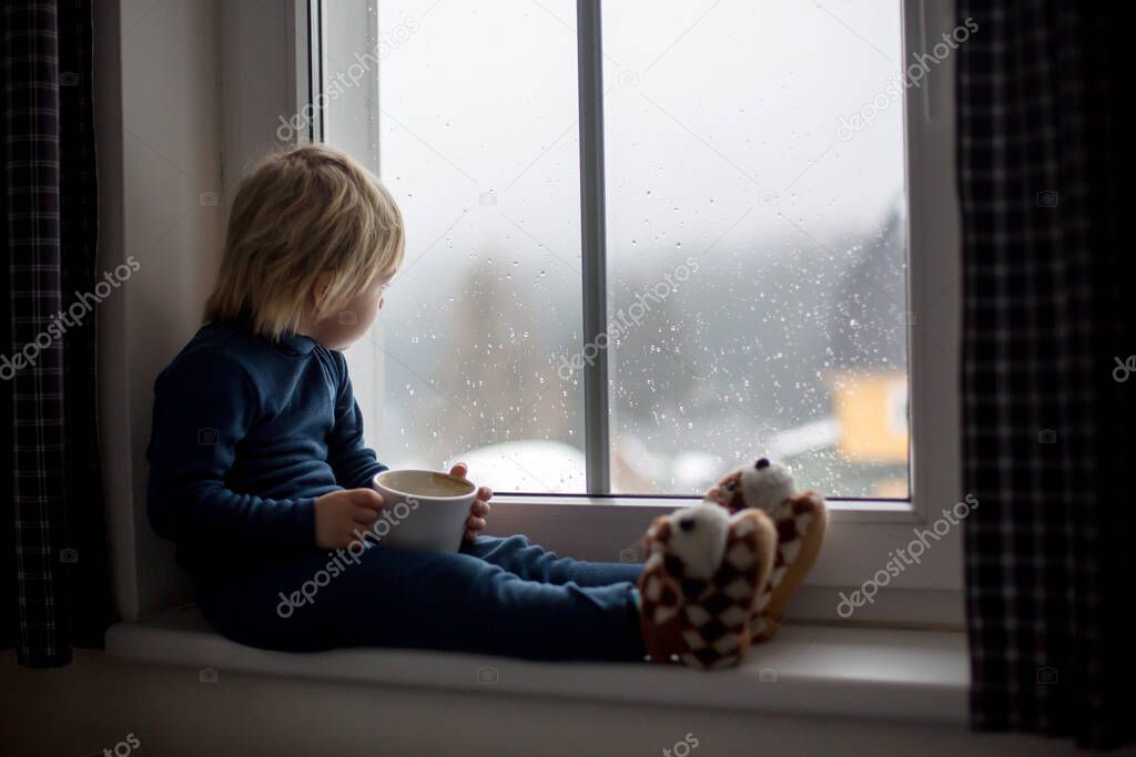 Toddler child, sitting on the window, watching the snow falling, enjoying cup of tea