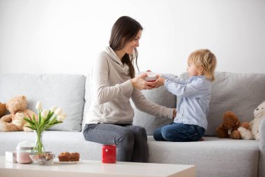 Güzel sarışın çocuk, güneşli oturma odasında anneler günü için küçük hediyeler vererek annesine çiçek ve kutu veriyor.