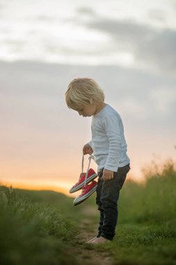 Mutlu çocuk, elinde bir çift spor ayakkabı, kırsal bir yolda yürüyor, çıplak ayakla.