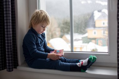Küçük çocuk, pencerede oturuyor, karın düşüşünü izliyor, küçük kitap okuyor.
