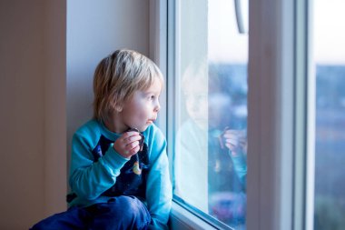 Küçük çocuk, gün batımında pencerede oturuyor, dışarı bakıyor.