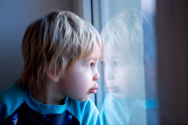 Küçük çocuk, gün batımında pencerede oturuyor, dışarı bakıyor.