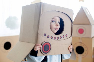 Tatlı çocuk, astronot gibi giyinmiş, evde karton roket ve kutudan yapılmış el yapımı kaskla oynuyor.
