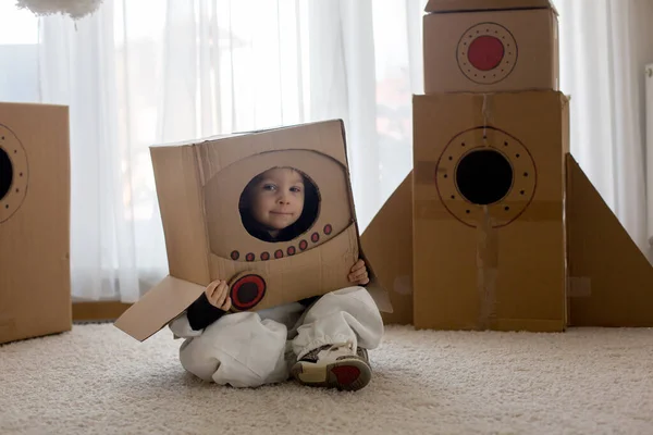Sweet toddler boy, dressed as an astronaut, playing at home with cardboard rocket and handmade helmet from box