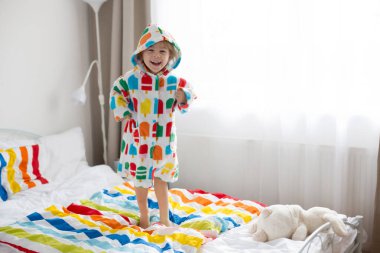 Mutlu çocuk, renkli bornozlu sarışın çocuk, yatağa atlıyor, mutlu bir şekilde gülümsüyor.