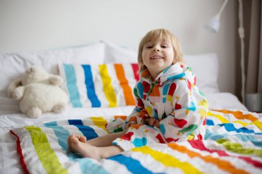 Mutlu çocuk, renkli bornozlu sarışın çocuk, içi doldurulmuş oyuncakla banyodan sonra yatakta oturuyor, gülümsüyor, renkli bir yatak.