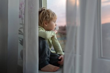 Sarışın çocuk, gün batımında pencerede oturuyor, dışarı bakıyor.