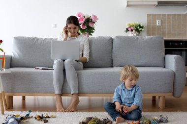 Anne, dizüstü bilgisayarı üzerinde çalışıyor ve telefonlara bakıyor. Annesi evde çalışırken yanında oynayan bir çocuk.