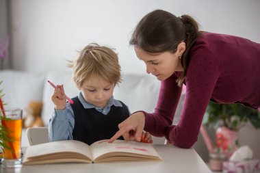 Sevimli bir anaokulu çocuğu, sarışın bir çocuk, bir iş kitabı ve boyama ödevi dolduruyor, annesi evde ona yardım ediyor.
