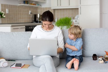 Anne, dizüstü bilgisayarı üzerinde çalışıyor ve telefonlara bakıyor. Annesi evde çalışırken yanında oynayan bir çocuk.