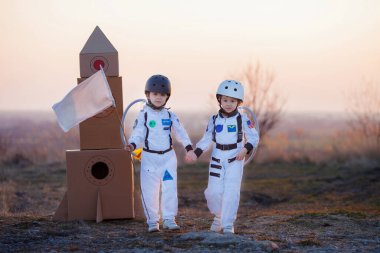 İki sevimli çocuk, erkek kardeşler, gün batımında parkta oynuyorlar, astronot gibi giyinmişler, ayda uçtuklarını hayal ediyorlar.
