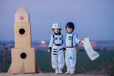 İki sevimli çocuk, erkek kardeşler, gün batımında parkta oynuyorlar, astronot gibi giyinmişler, ayda uçtuklarını hayal ediyorlar.